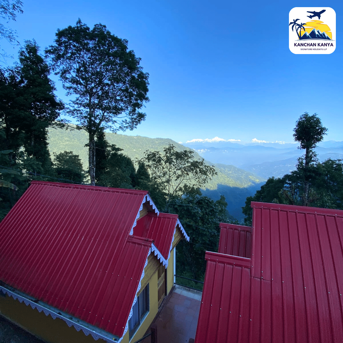Scenic hillside village with bright red roofs and tall trees, overlooking distant mountains under a clear blue sky; logo in the top-right corner.