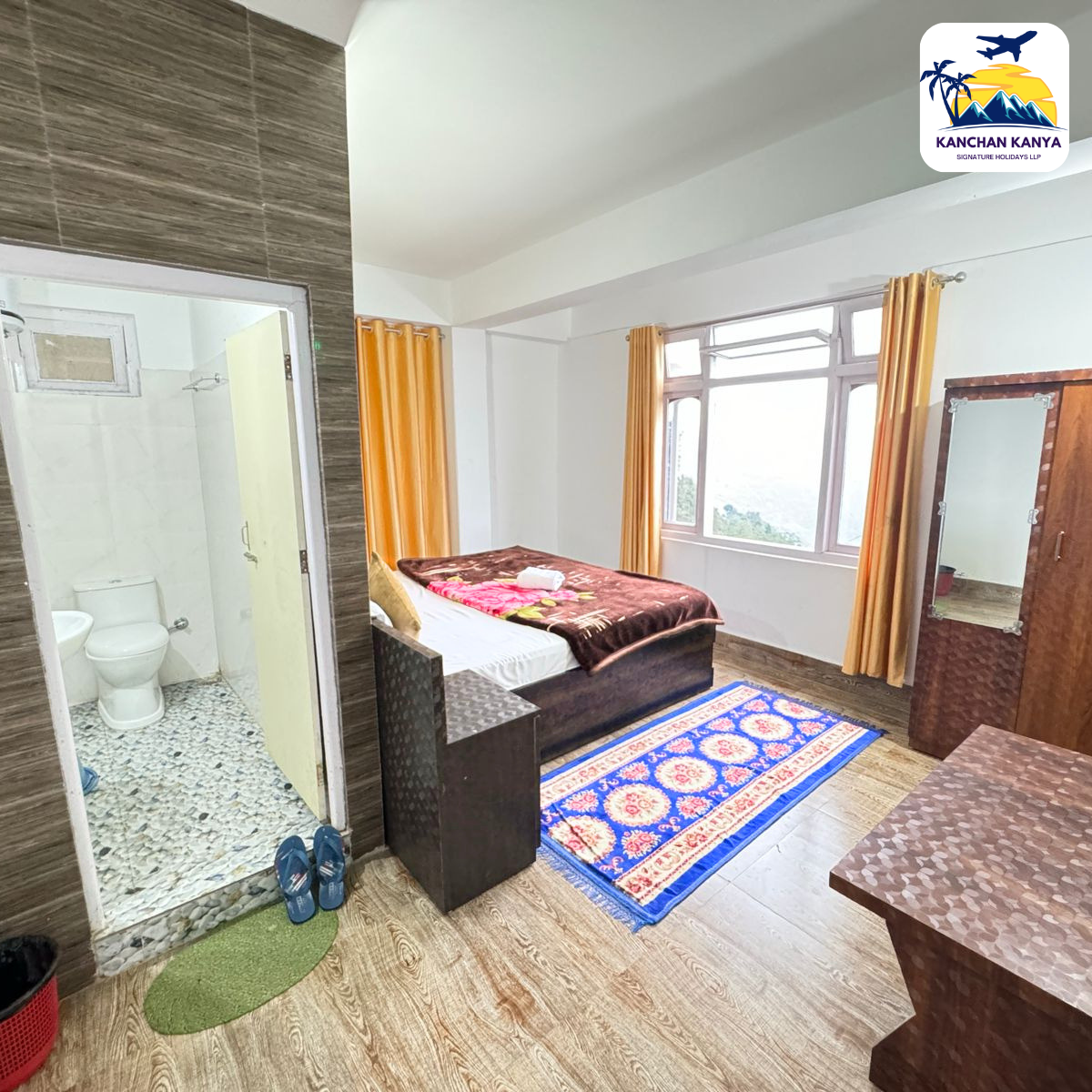 Hotel room with a double bed, brown bedding, yellow curtains, a wooden wardrobe with a mirror, and a blue patterned rug; slippers by the door and an open bathroom to the left.