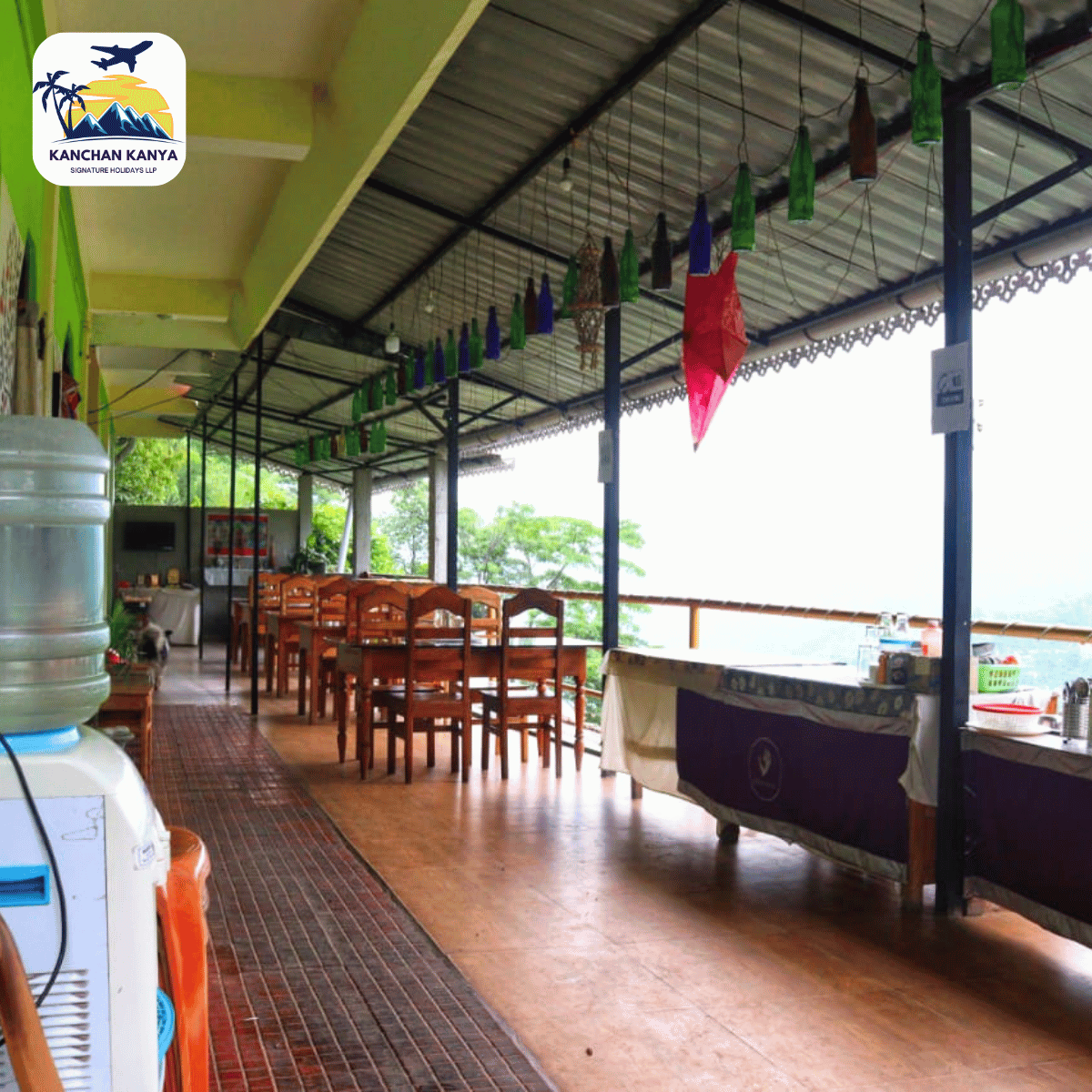 Open-air dining area with wooden tables and chairs under a roof, colorful hanging decorations, and a logo in the top-left corner.