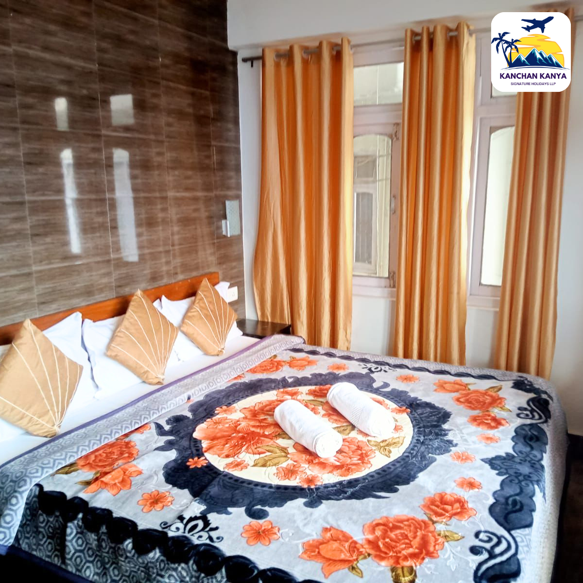 Double bed with orange‑pillow cushions and a floral throw blanket, two rolled white towels on top, beside orange curtains and a brown tiled wall.