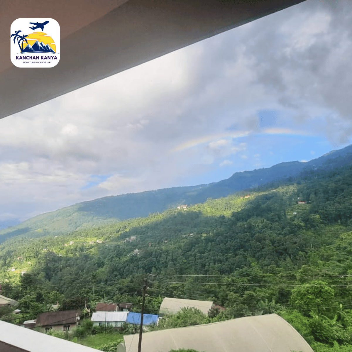 Green valley and forested mountains under a cloudy blue sky, seen from a balcony edge with a building roof in the foreground.