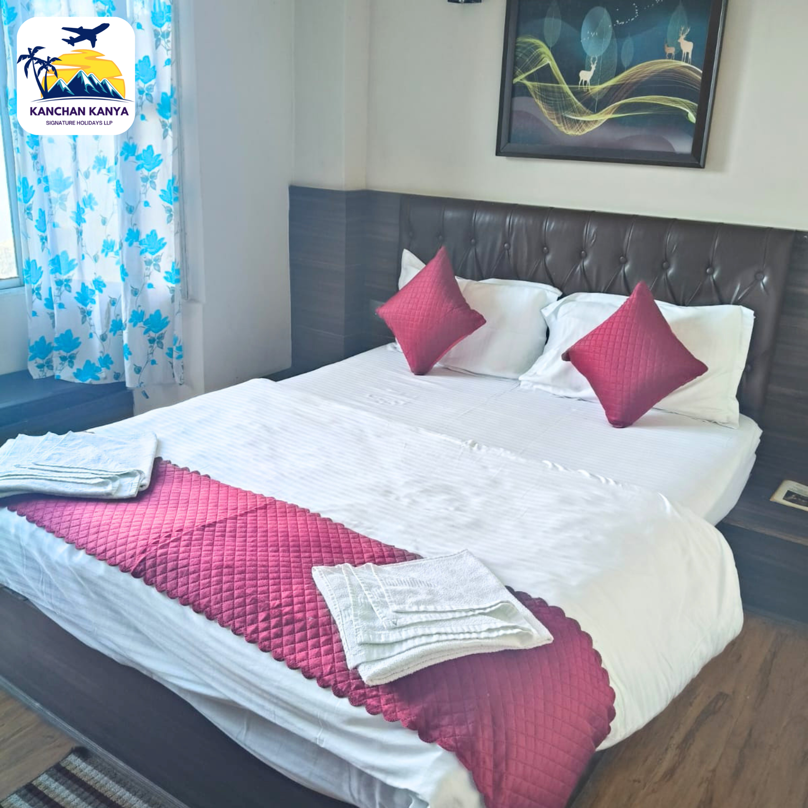 Hotel bedroom with a white bed, red quilted runners and pillows, folded towels, and a tufted headboard; blue floral curtains and wall art above the bed.