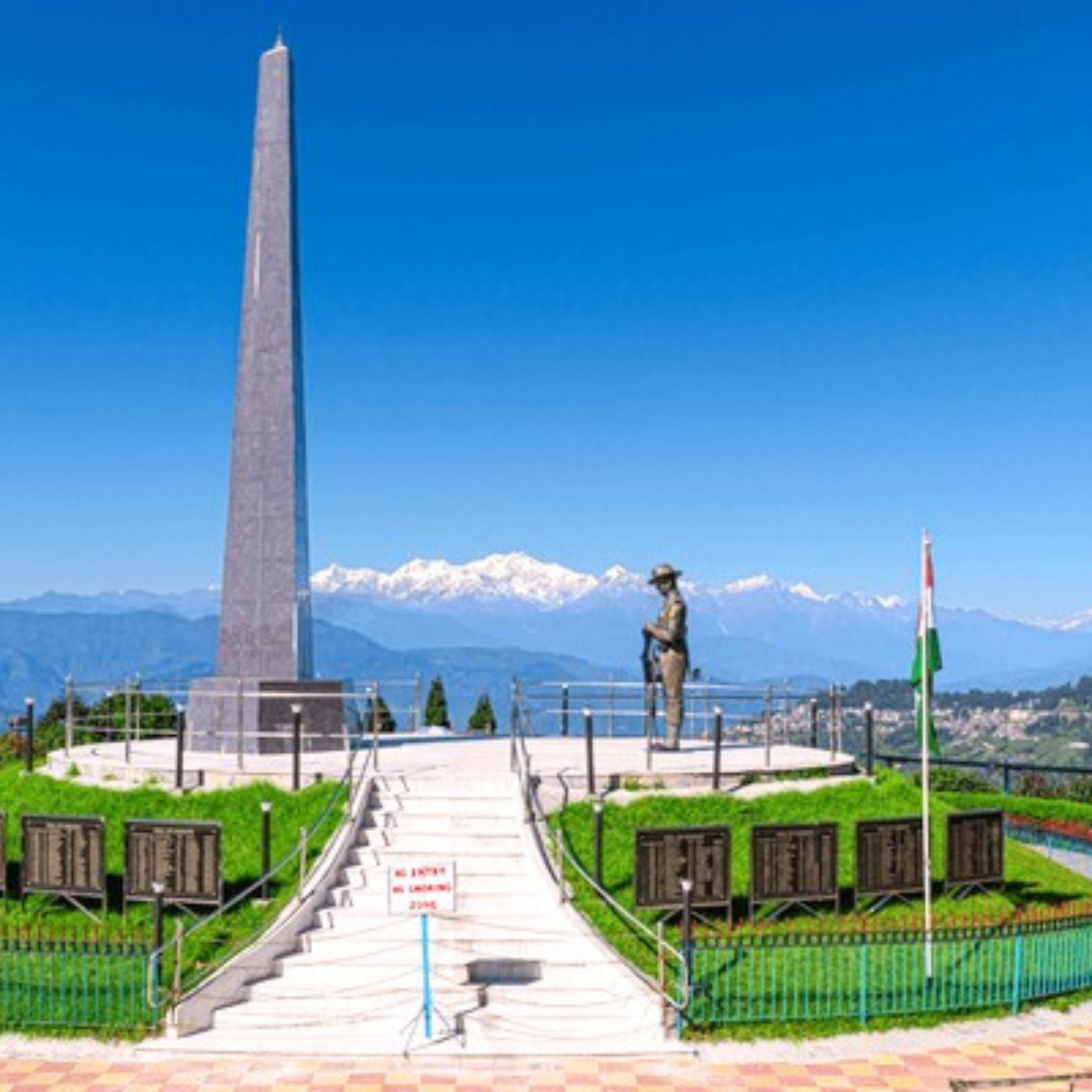 Tall obelisk memorial with a bronze soldier statue at the base, flanked by stairs and green railings against distant mountains and blue sky.