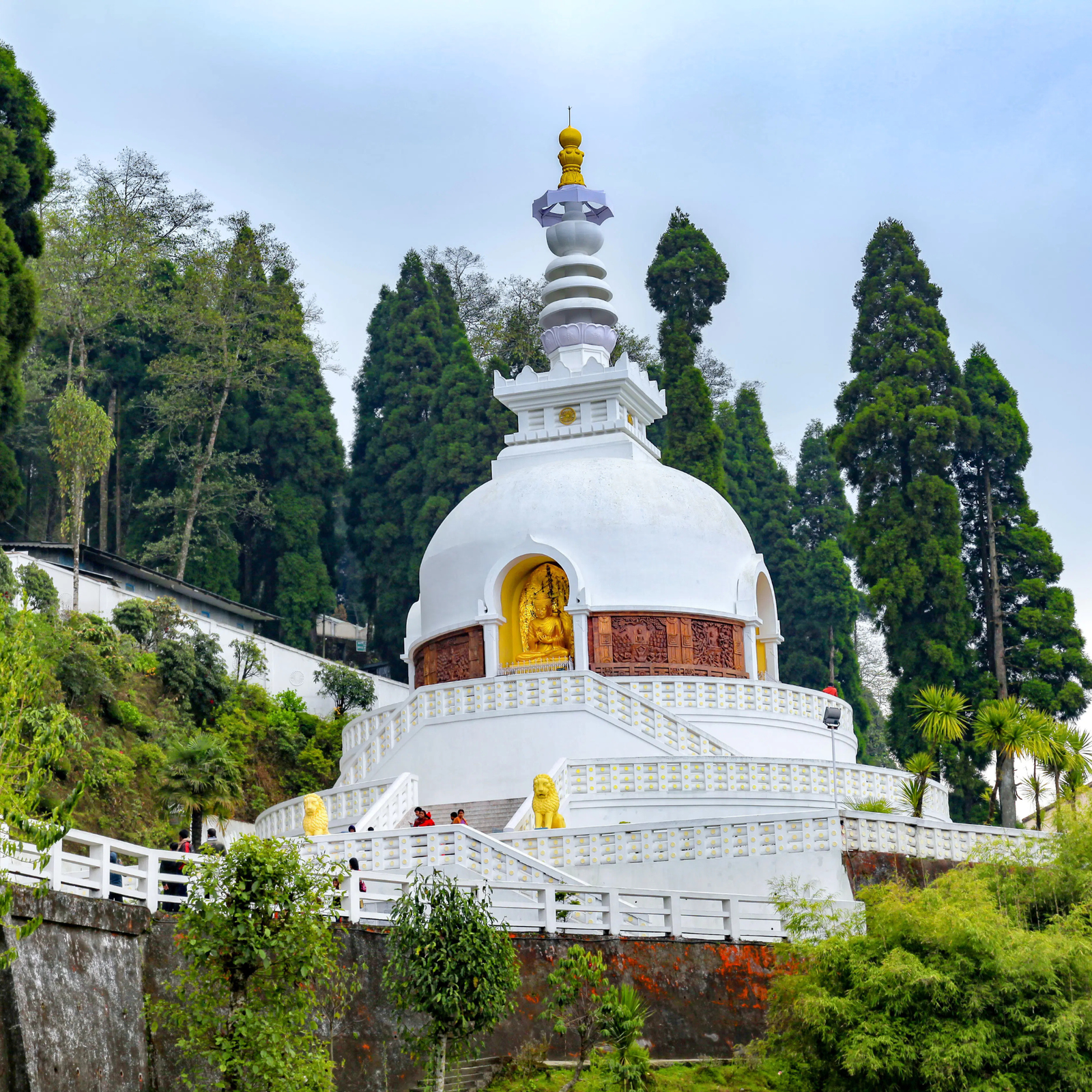White Buddhist stupa with a gold Buddha statue and a tall yellow spire, surrounded by trees and visitors on the steps.