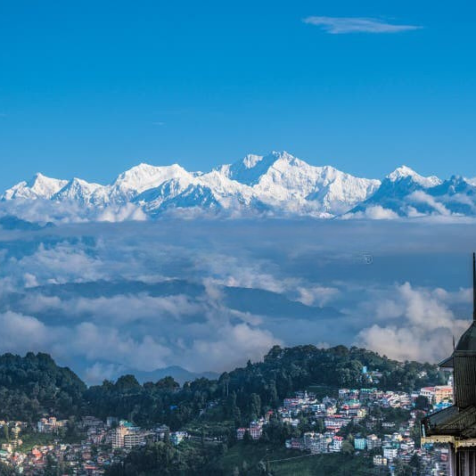 Snow-capped mountains overlook a town nestled in green hills under a clear blue sky with scattered clouds.