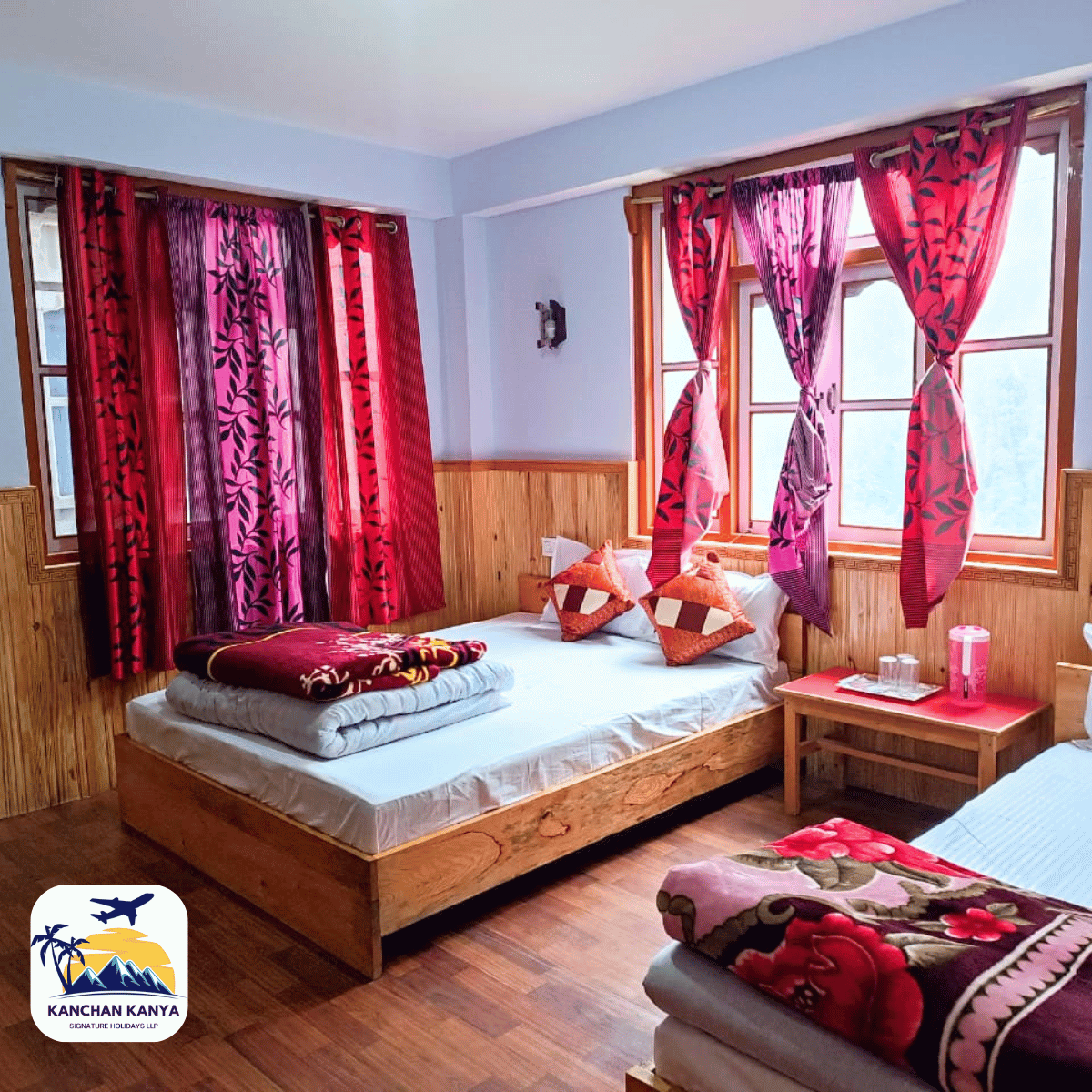 16 Cozy twin hotel room with wooden beds, two single beds, pink and purple patterned curtains, and a red bedside table with glassware; cushions and a folded blanket on the bed.