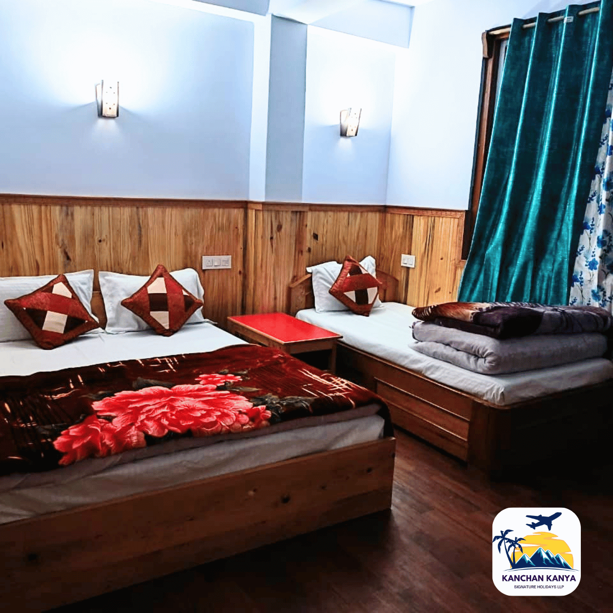 17 Hotel room with two twin beds, wooden paneling, blue walls, and teal curtains; blankets stacked on the right bed.