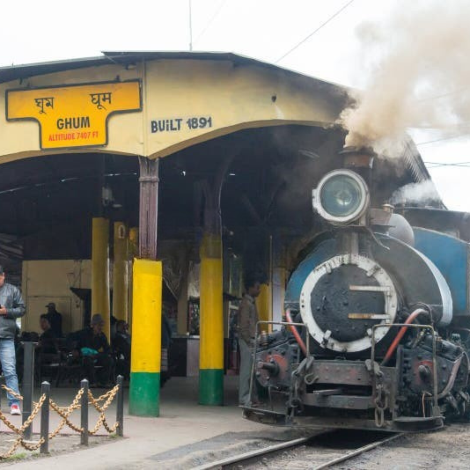 Old steam locomotive at a station, smoke puffing from the engine amid bright yellow pillar supports and tracks in view.