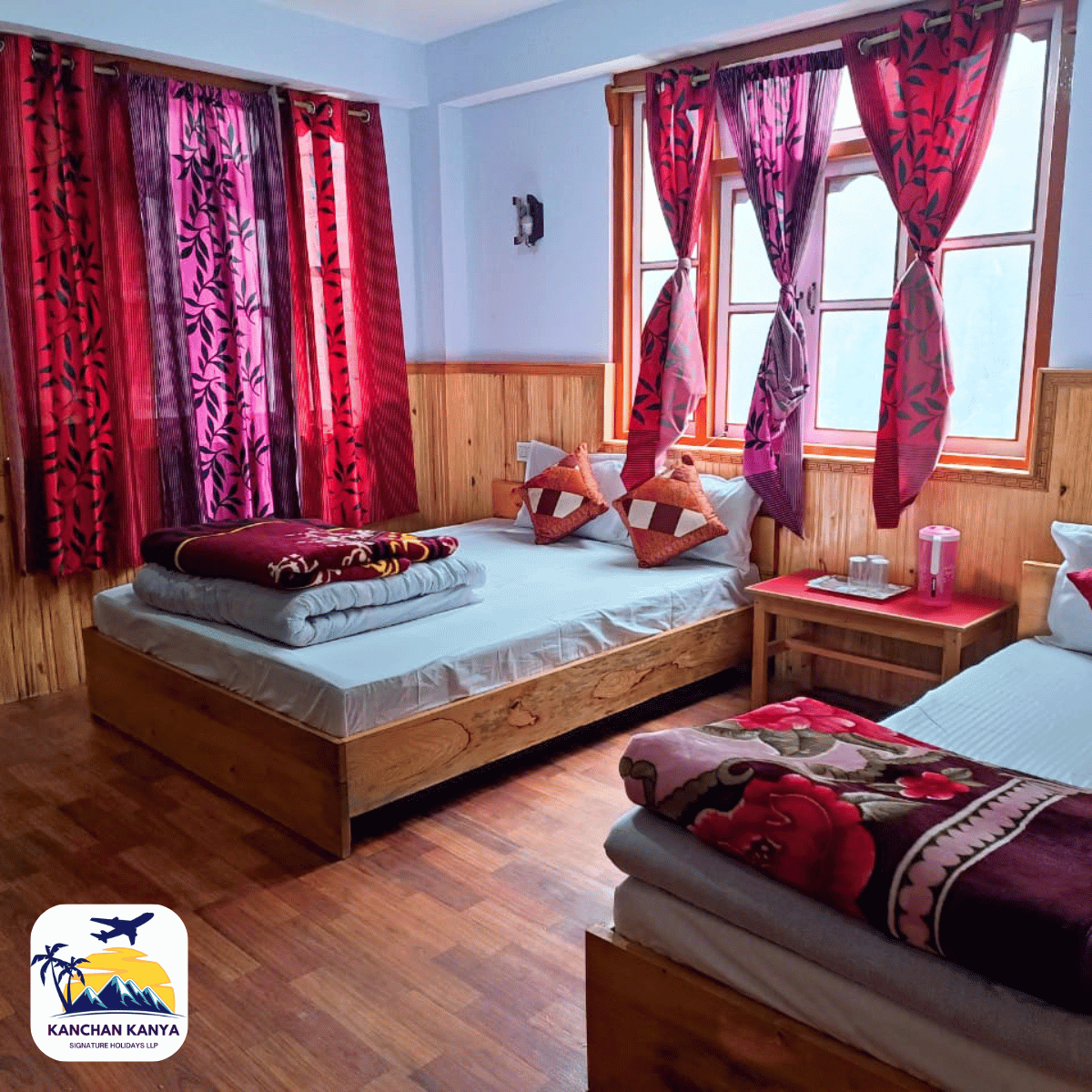 20 Two single beds with wooden frames in a blue-painted room, pink-purple curtains, and a small bedside table between them with a pink thermos and cups.