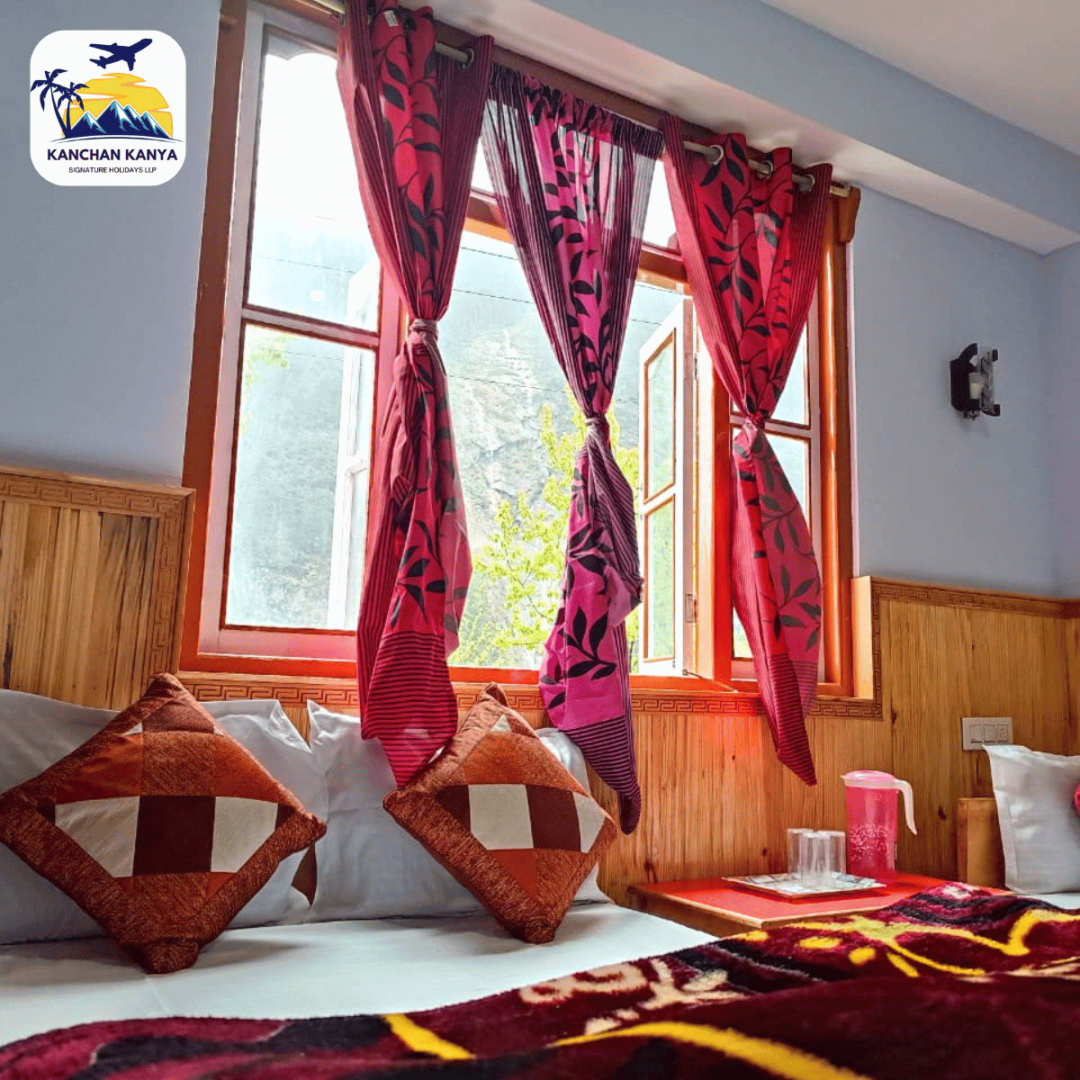 21 Hotel room interior with a wooden-framed window, pink patterned curtains, bed with brown diamond pillows, and a colorful blanket; logo in the top-left corner.