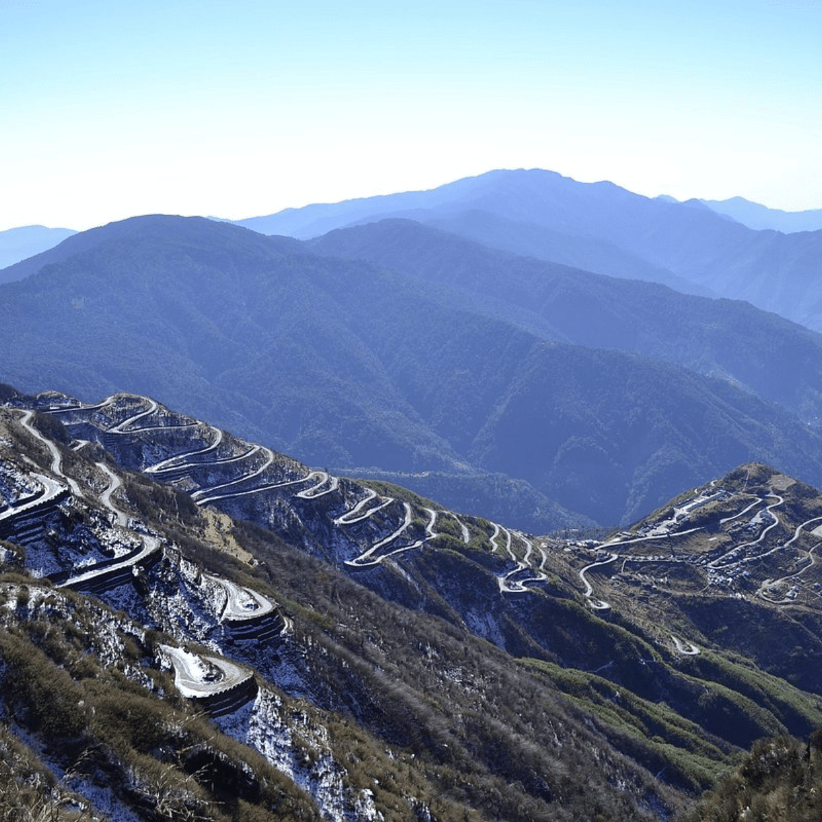 24 Winding switchback road climbs a snow-dusted mountain slope beneath a blue sky.