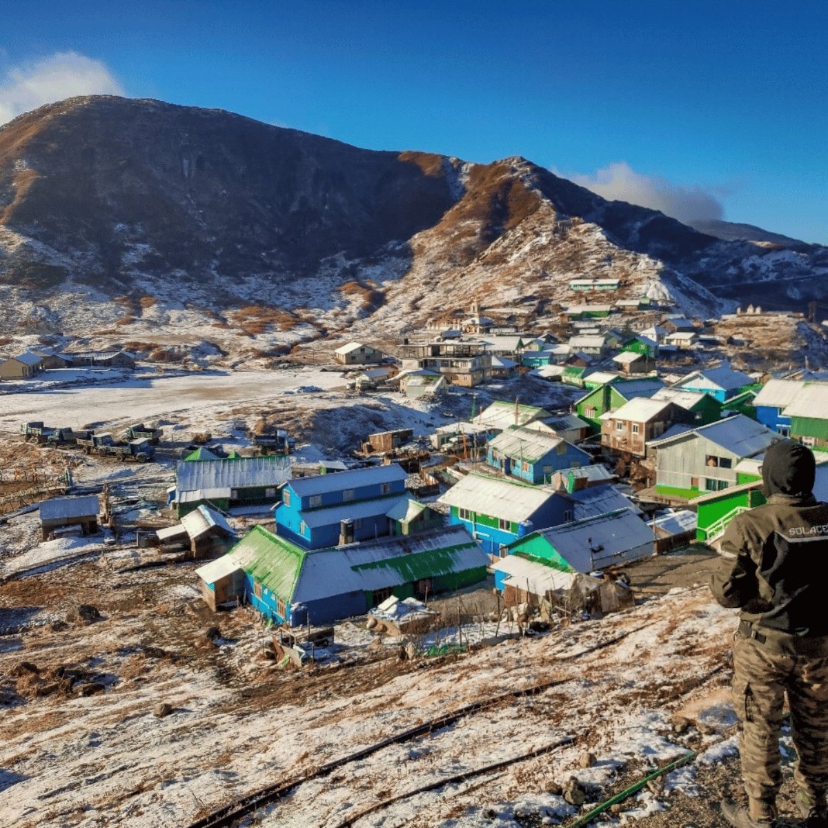 25 Snowy mountain village with green and blue houses nestled on a slope, a hiker in the foreground observes the scene.