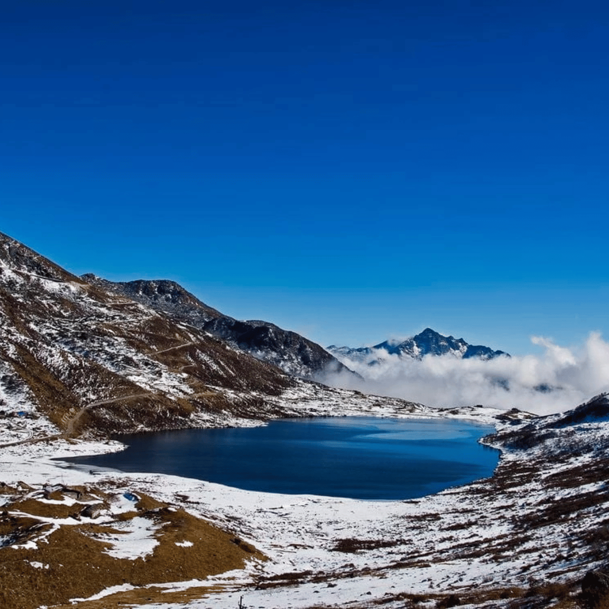 26 Snow-dusted mountains circle a dark blue alpine lake under a clear blue sky with distant clouds on the horizon.