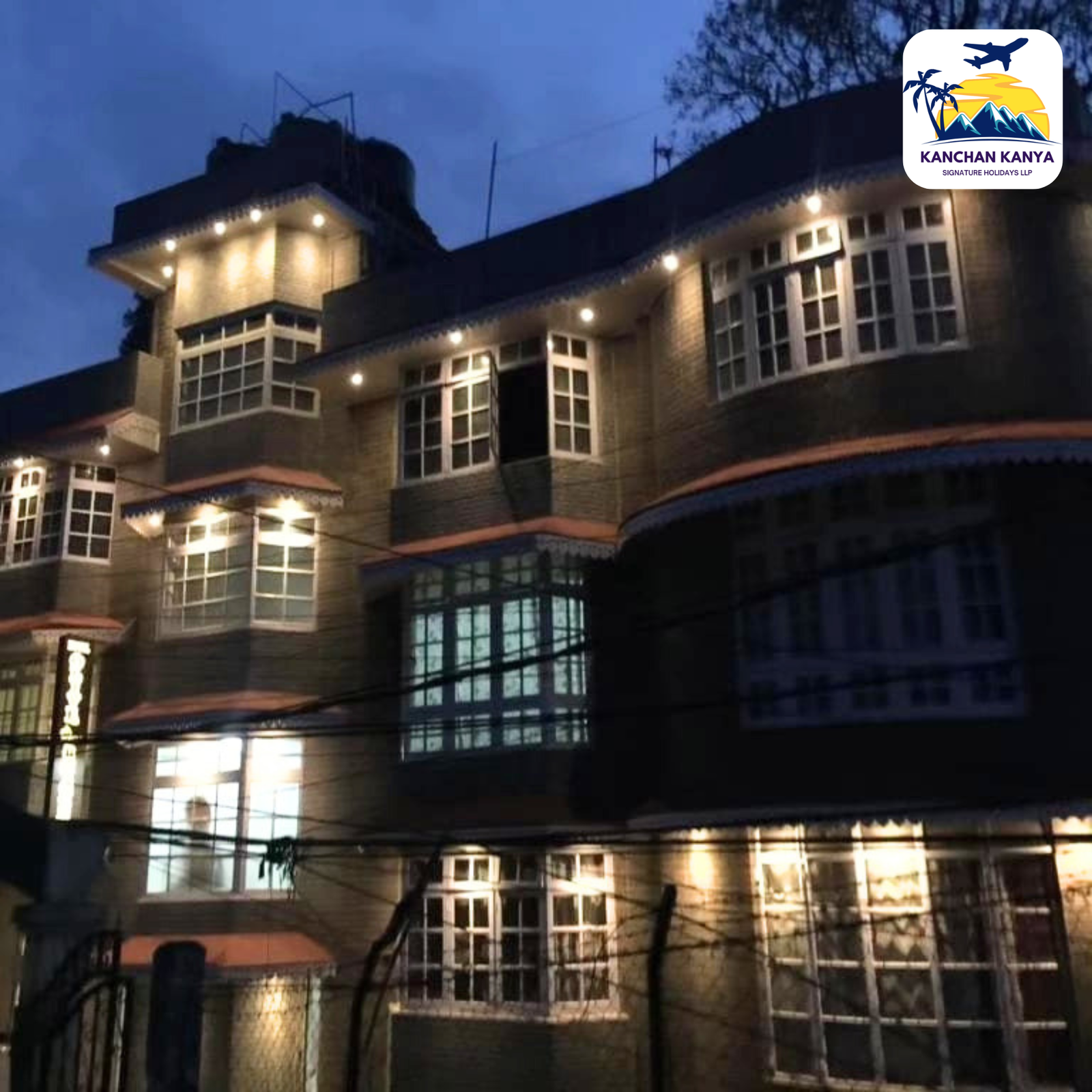 Night view of a brick building with illuminated windows and string lights; logo of Kanchan Kanya in the top-right corner.