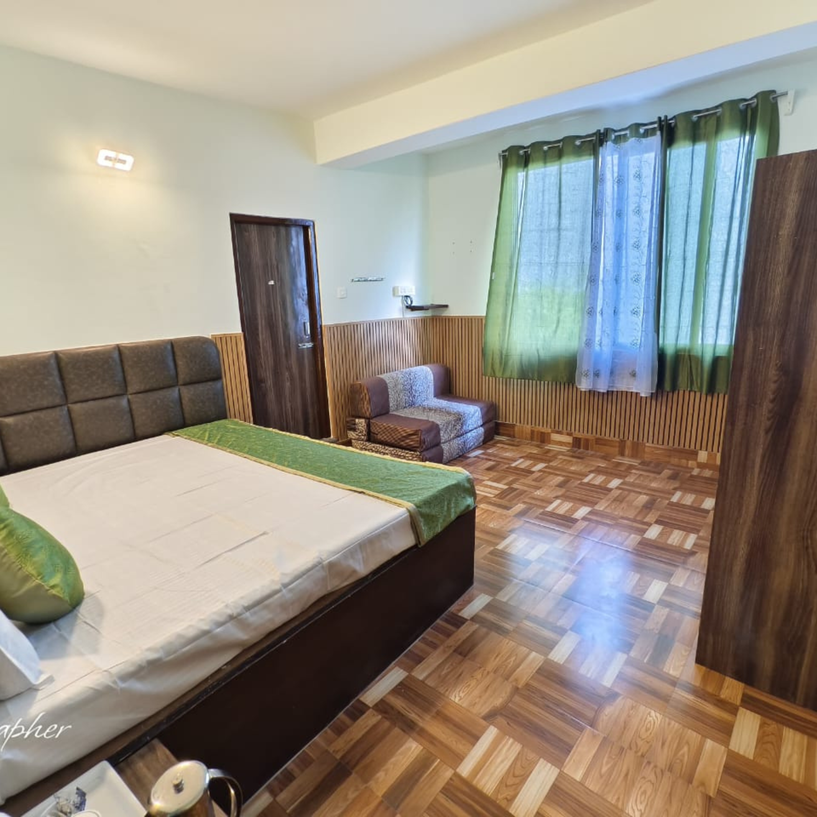 Hotel room with a large bed, brown upholstered headboard, wooden paneling, and a window with green sheer curtains.