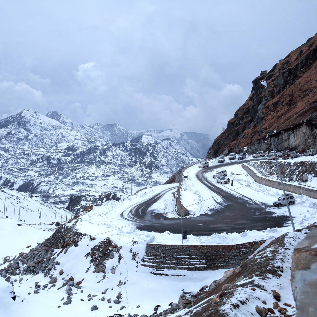 35 Snow-covered alpine road winding along a rocky hillside with cars parked along the curve and snowy ground.