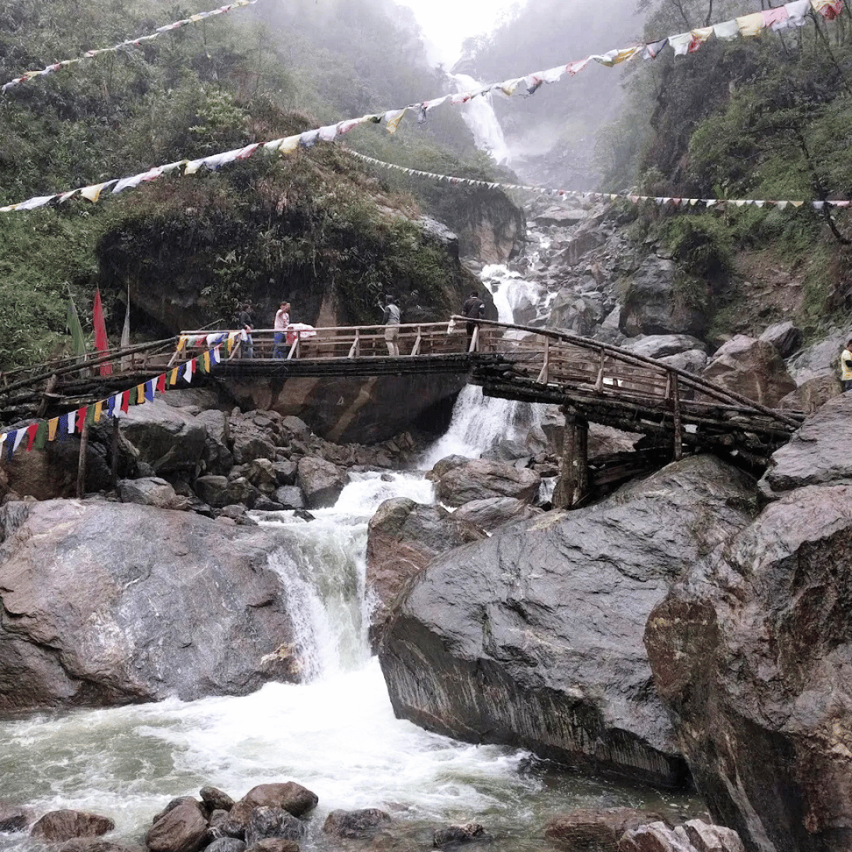 37 Wooden footbridge across a rocky mountain river with a distant waterfall; colorful prayer flags hang above and people stand on the bridge in a misty canyon.