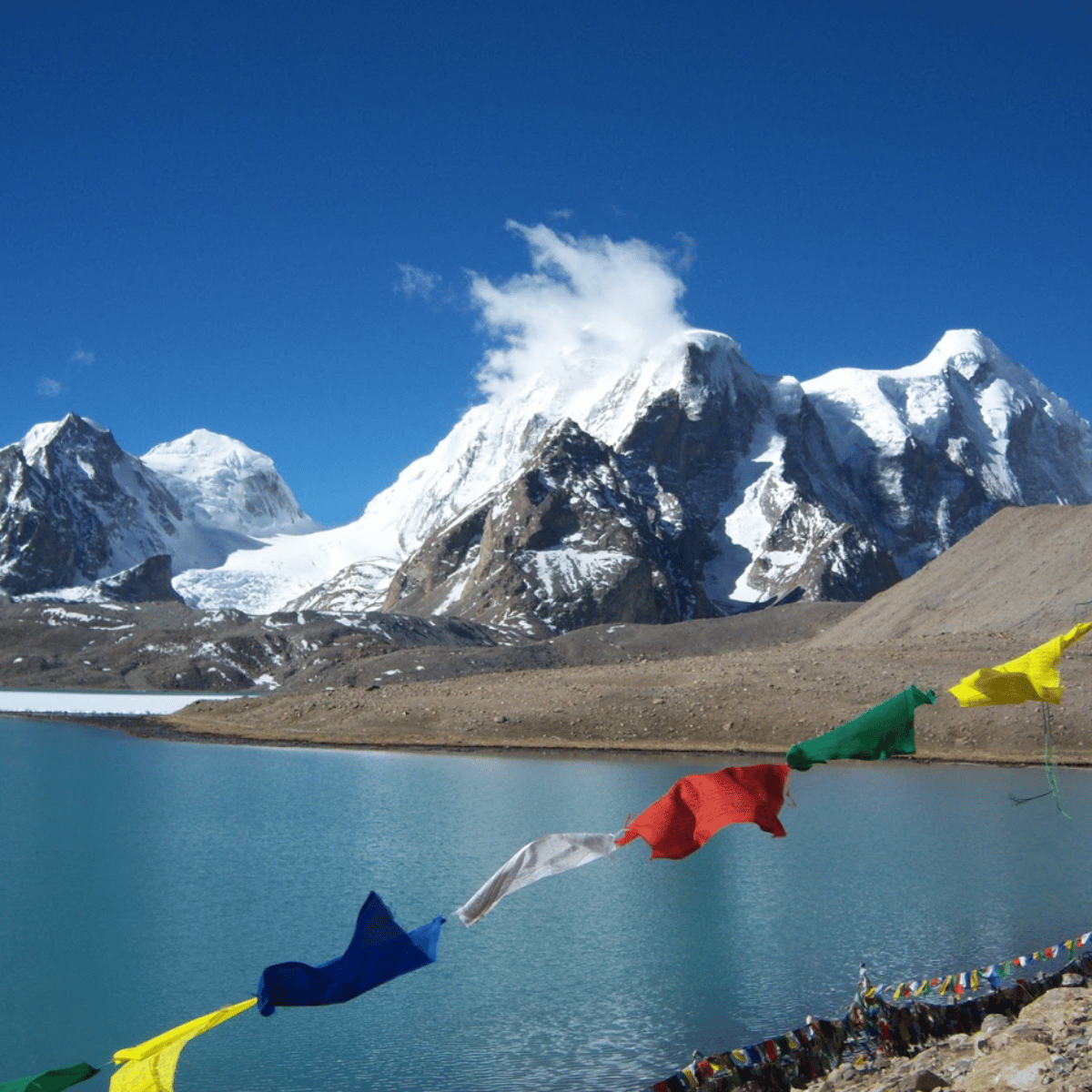 38 Snowy mountain range rises behind a turquoise lake, with colorful prayer flags fluttering along the shore.