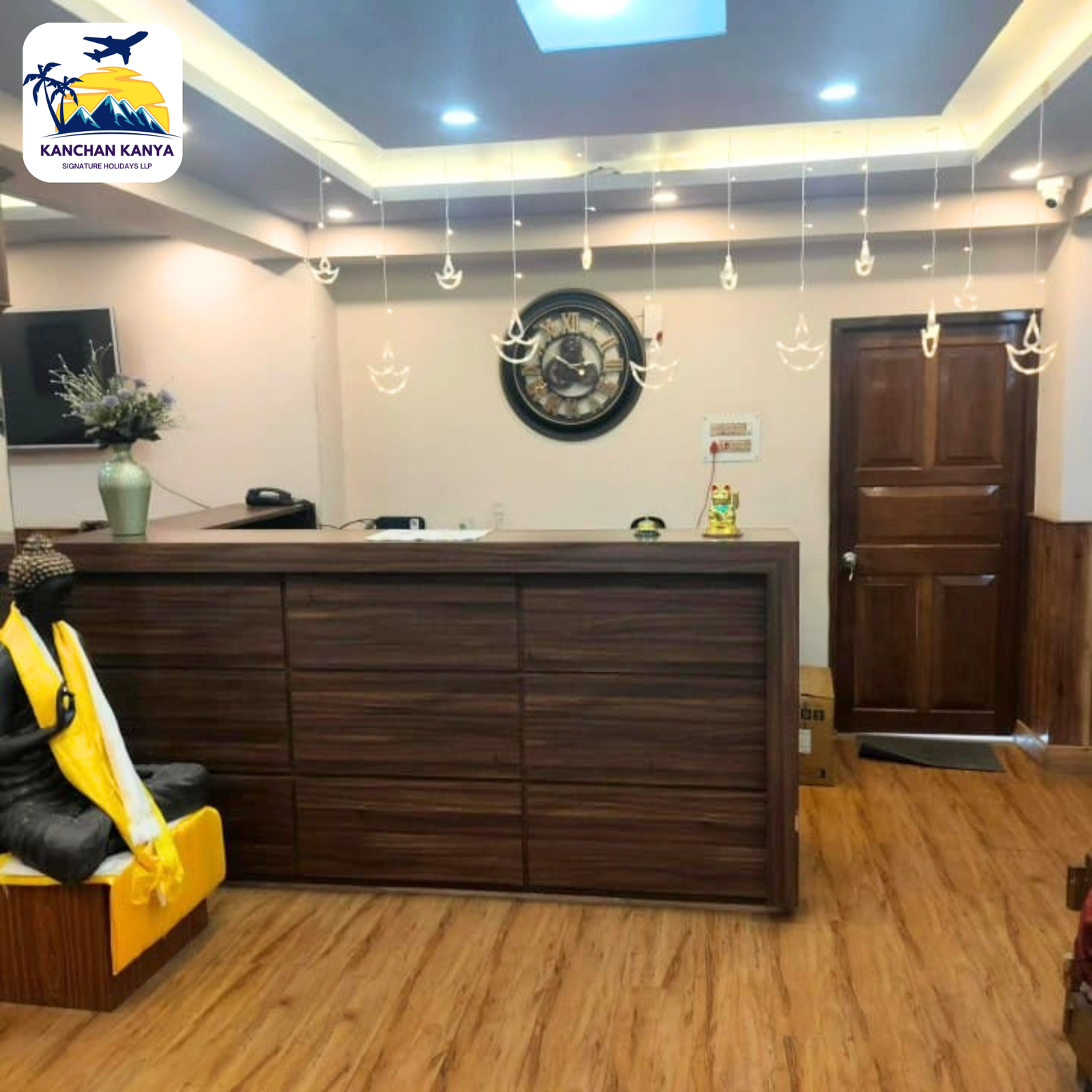 Reception desk in a hotel lobby with a large wall clock and hanging decor; logo in top-left corner reads 'KANCHAN KANYA'.