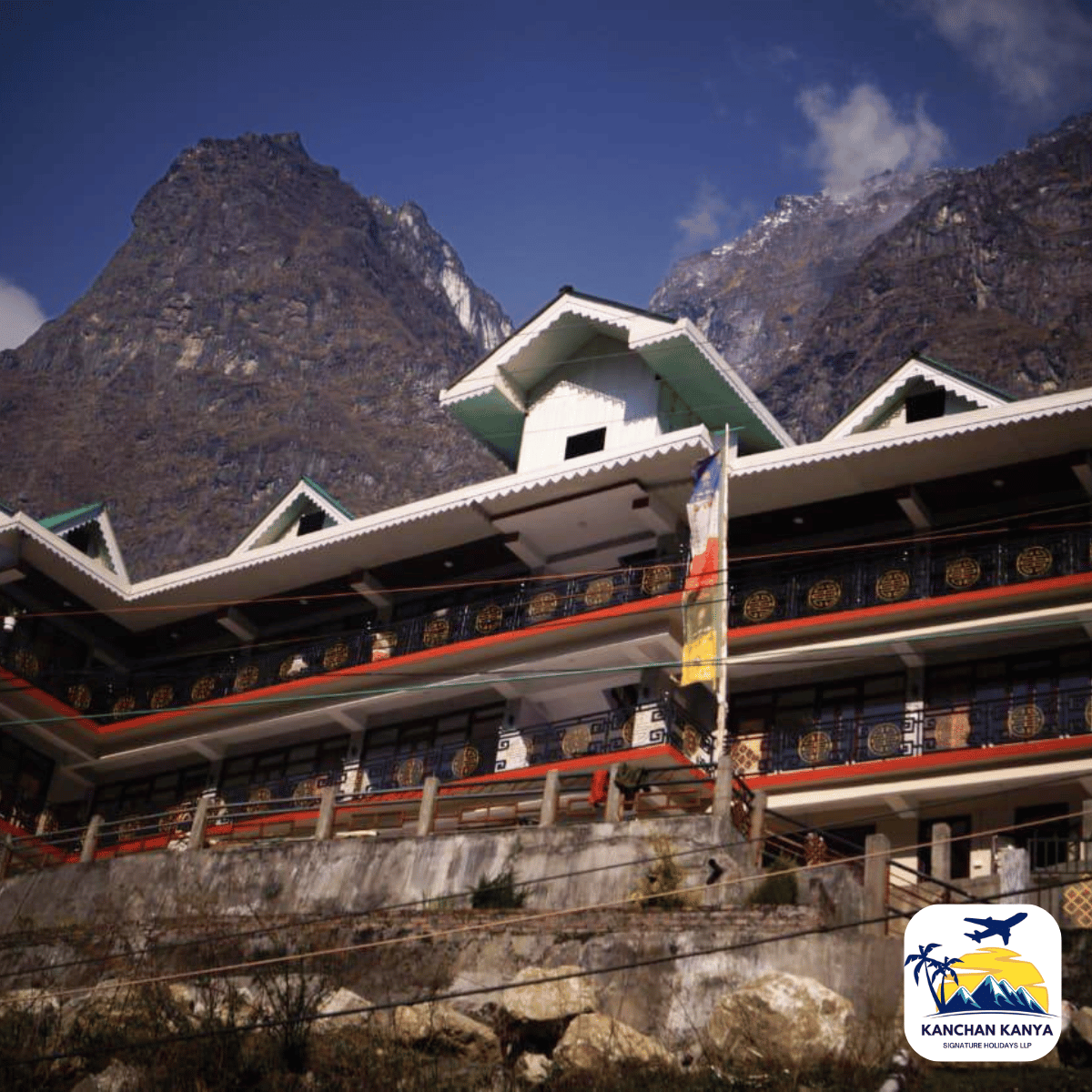 4 Three-story hillside hotel with decorative white-green roofs, set against rugged mountains under a blue sky, logo bottom-right corner of the image.