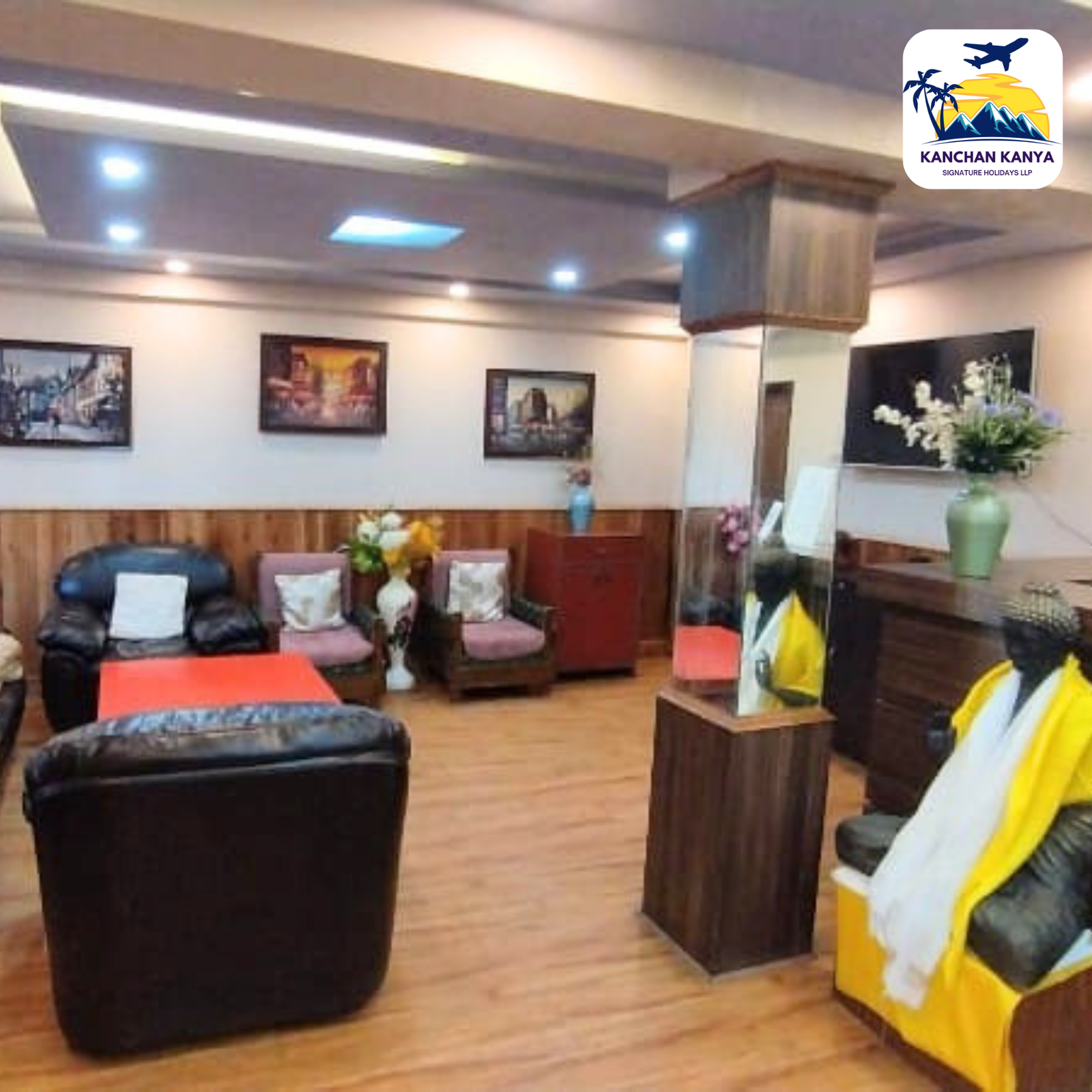 Hotel lobby with brown leather seating, wooden tables, wall art, and a large brand logo in the top-right corner.