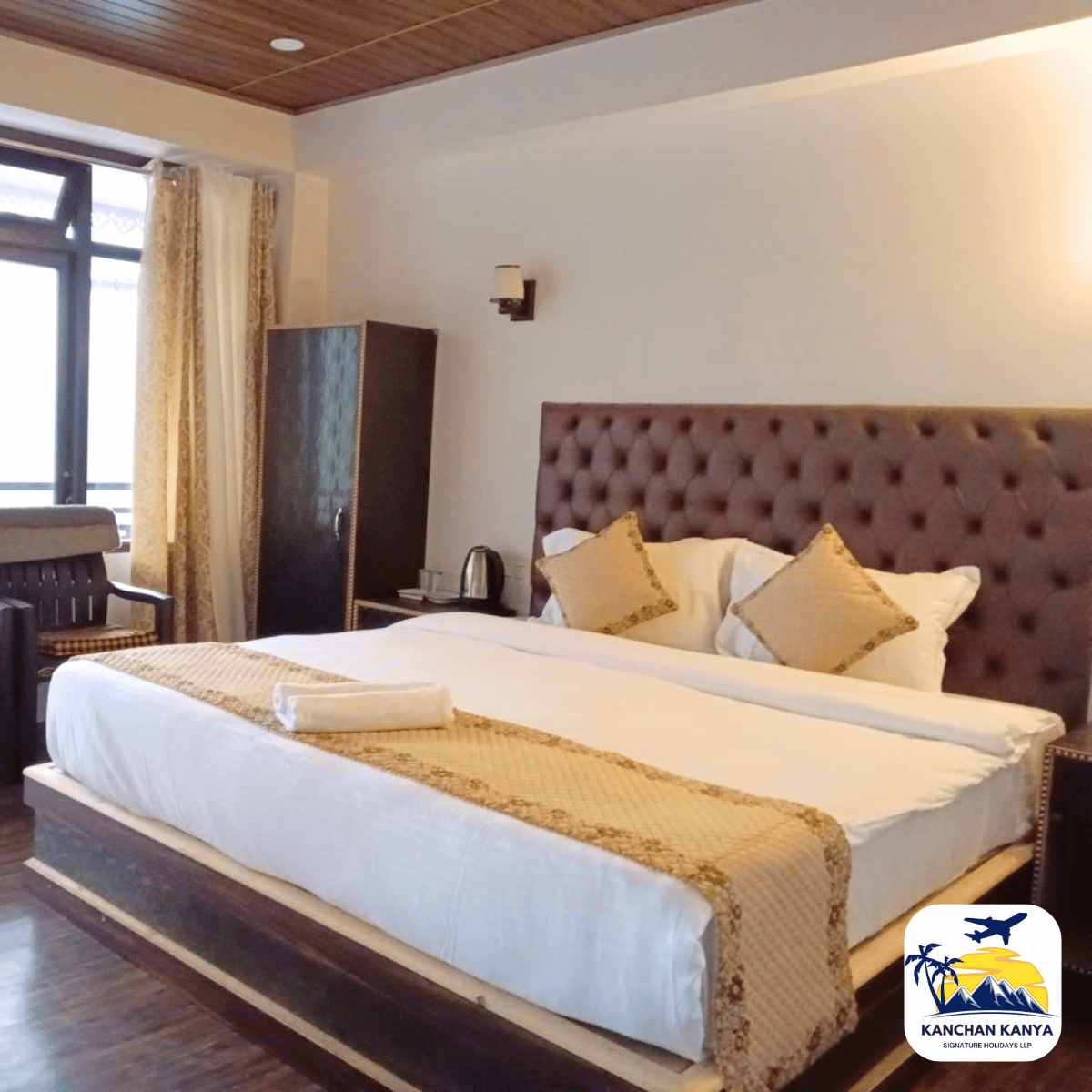 5 Hotel room with a large bed and tufted brown headboard, beige bedding, decorative cushions, and a window with curtains beside a wardrobe.