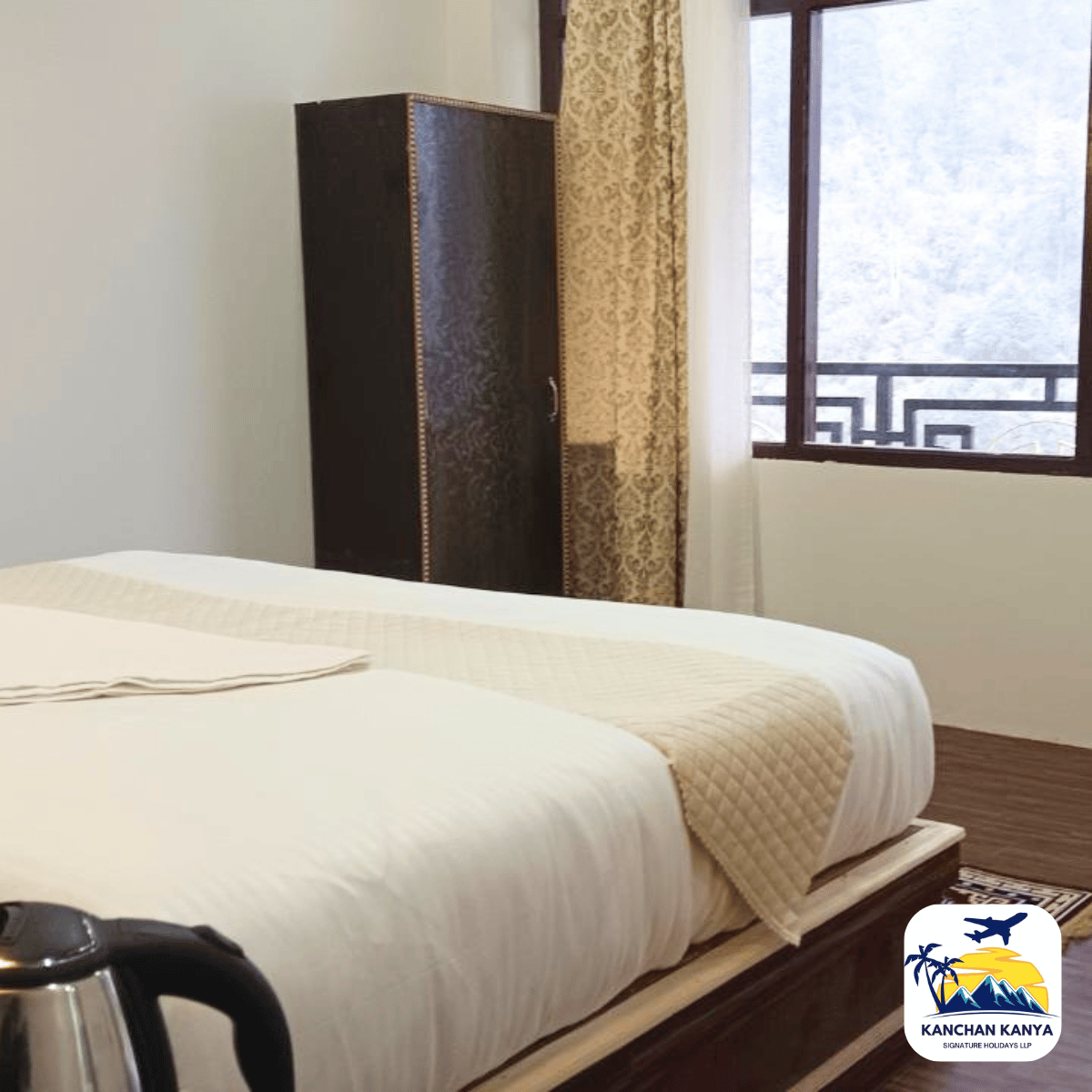 6 Hotel room featuring a neatly made bed with white linens and a beige quilt, a dark wardrobe, patterned curtains, and a window with decorative railing; an electric kettle sits on a table in the foreground; logo bottom right.