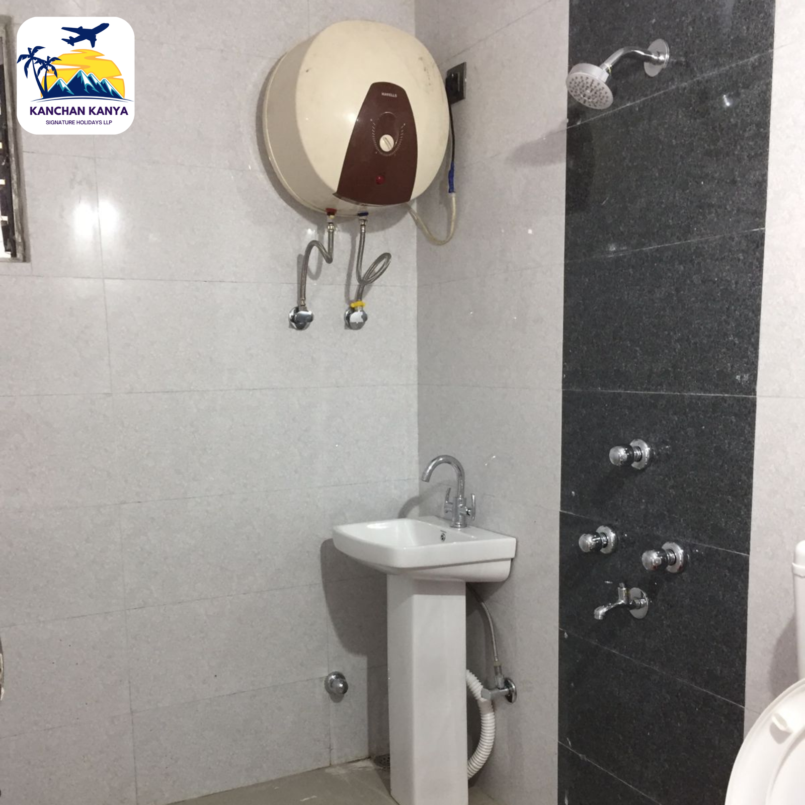 Small bathroom showing a wall-mounted sink with plumbing and a round water heater on light tiles; a shower with multiple controls on a dark tile wall, and a logo in the top-left corner reading 'KANCHAN KANYA'