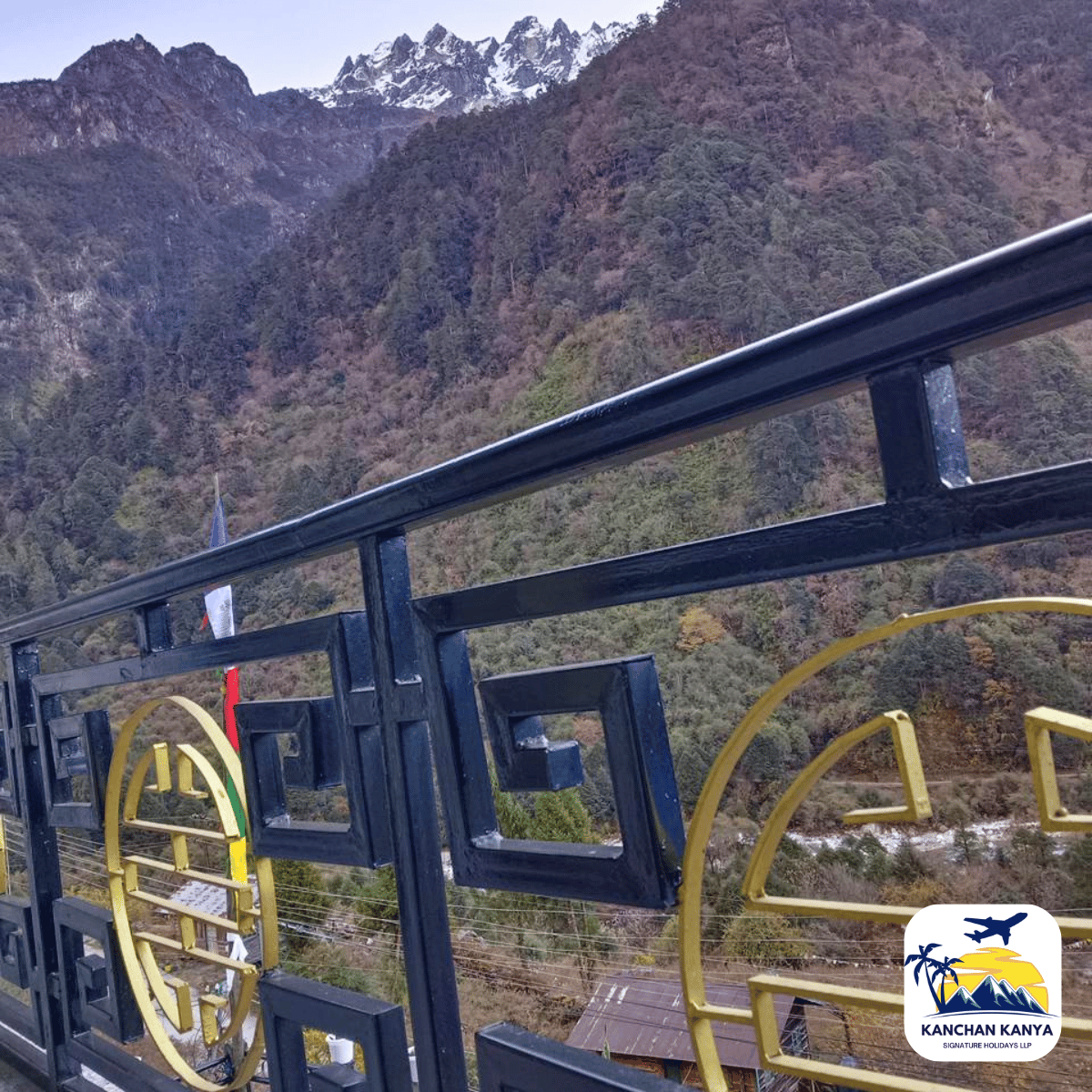 9 Blue metal railing with geometric shapes overlooks a forested mountain landscape with snow-capped peaks.
