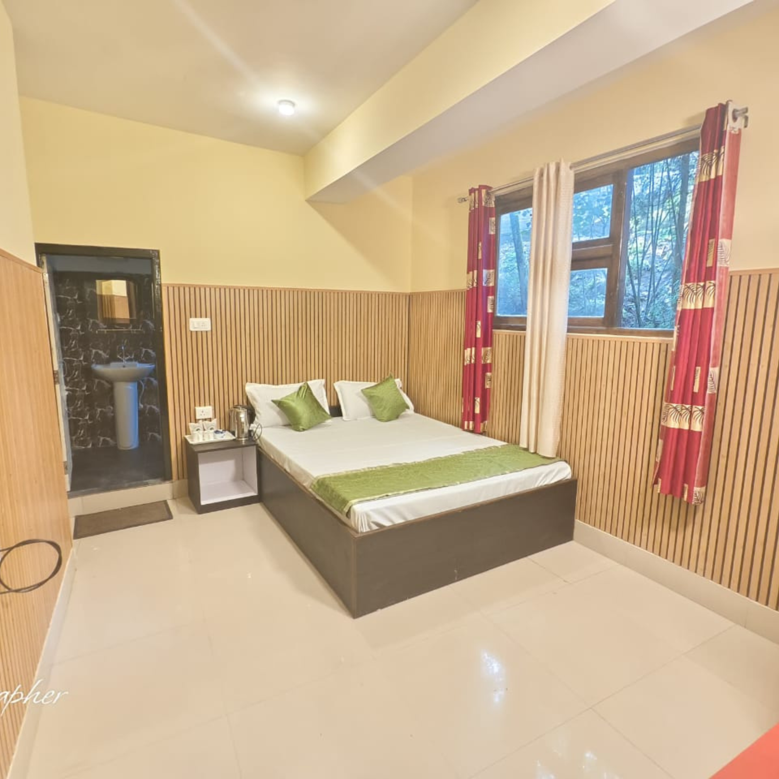 Hotel-style bedroom with a platform bed, white linens and green pillows, wood-paneled walls, and a large window with red curtains.
