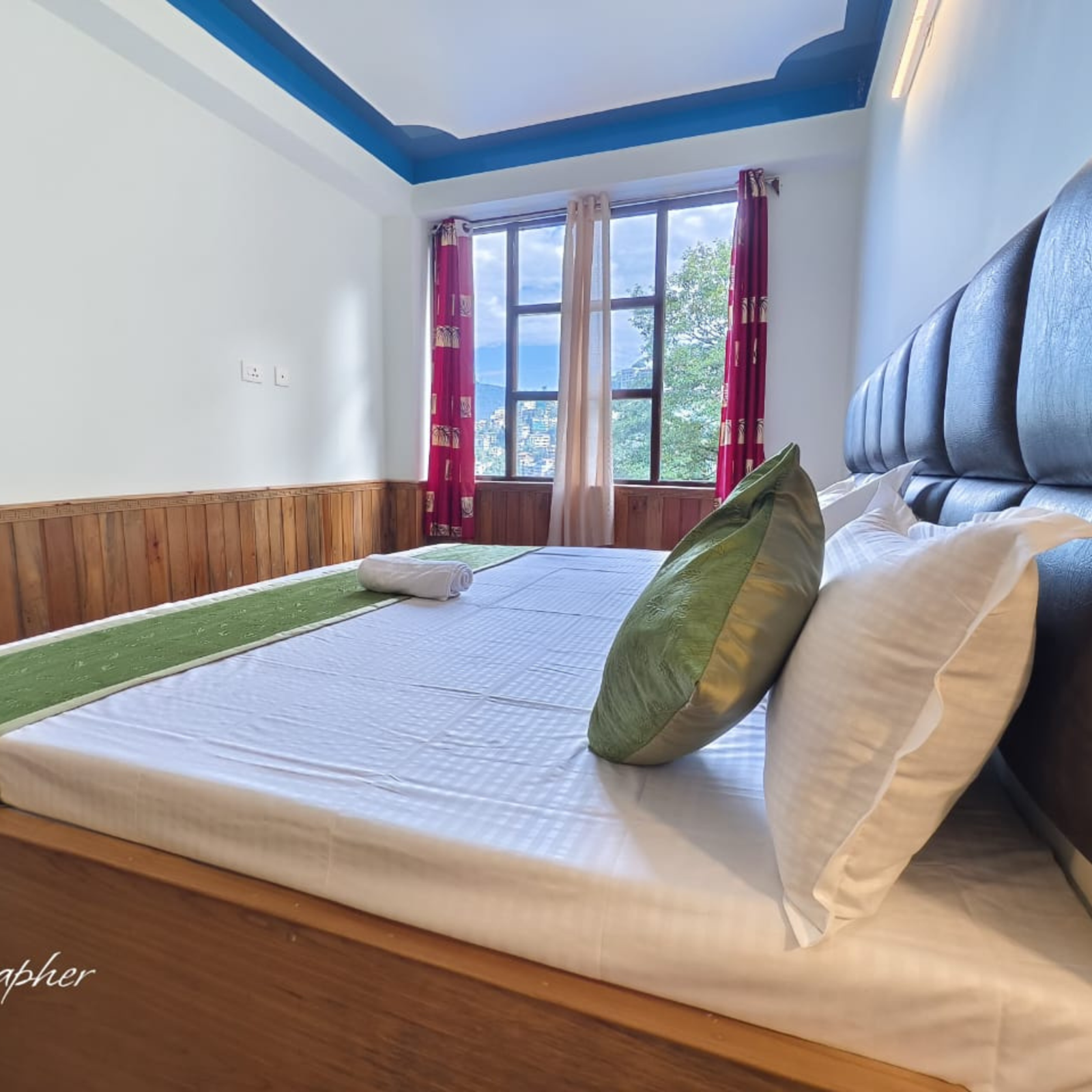 Neatly made bed with green pillows in a bright room, large window with red curtains and a mountain view outside, a rolled towel on the bed.