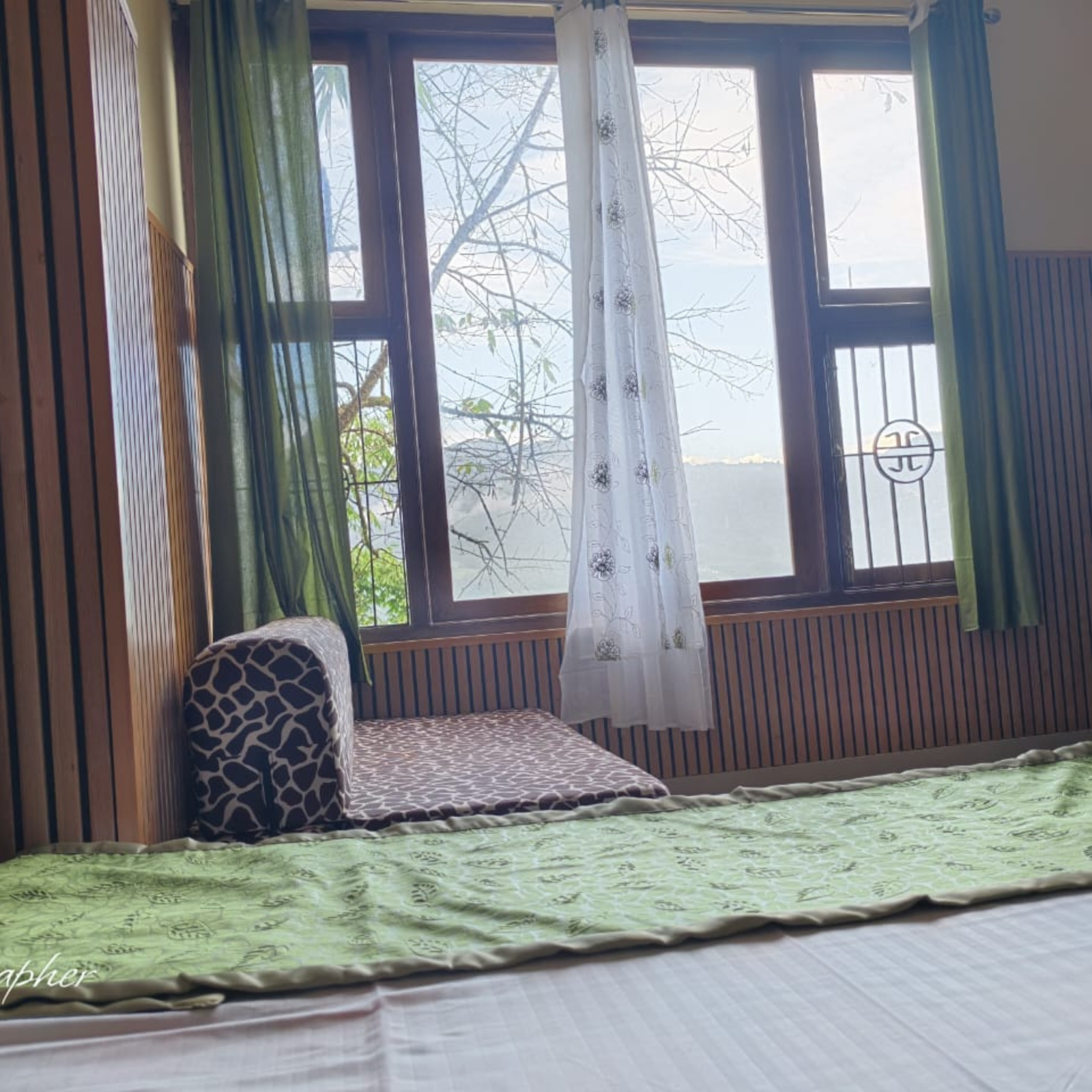 4 Bedroom corner with a large window, green and white curtains, and wooden paneled walls framing a view of leafless tree branches outside.