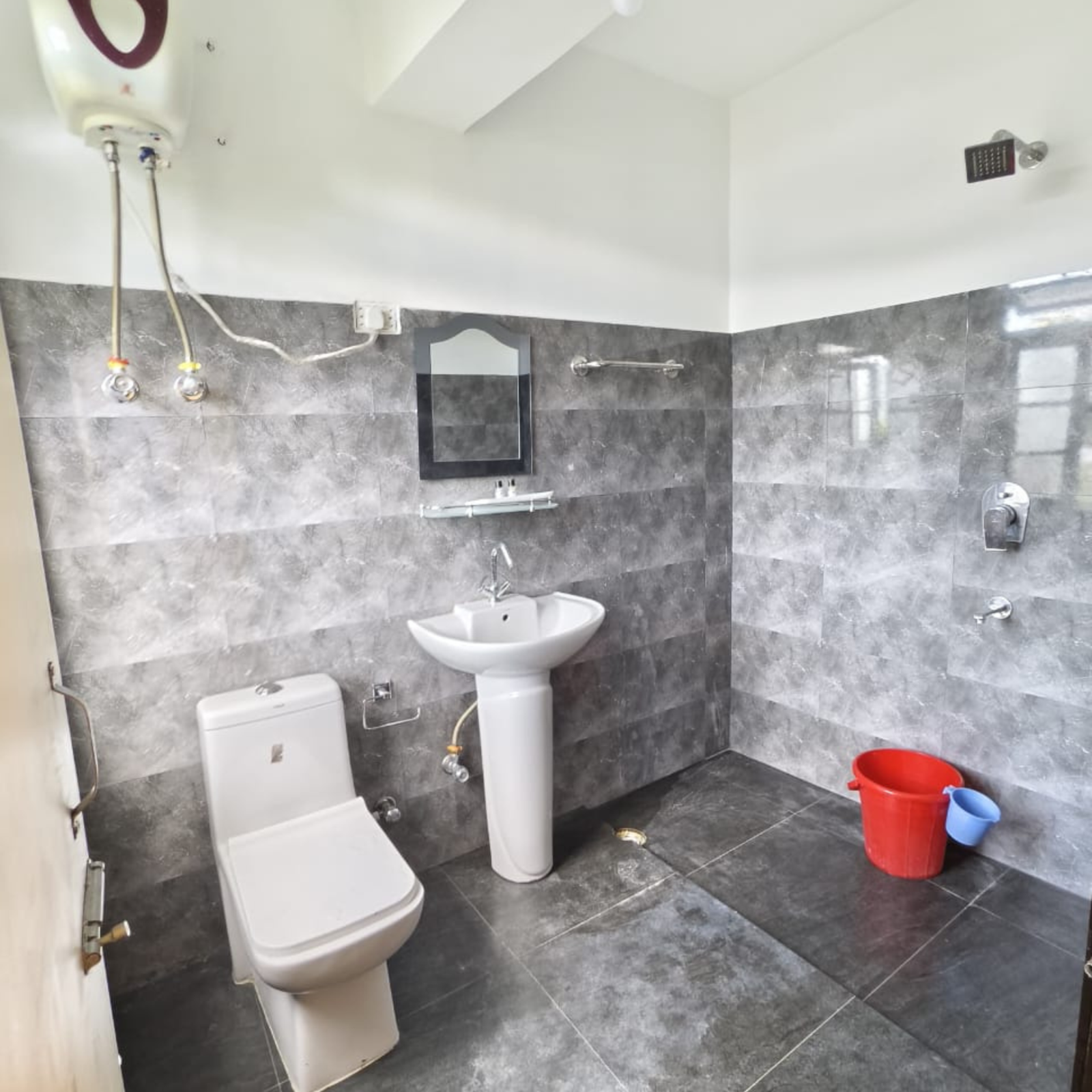 Gray-tiled bathroom with a white toilet and pedestal sink, wall mirror, and red and blue buckets on the floor