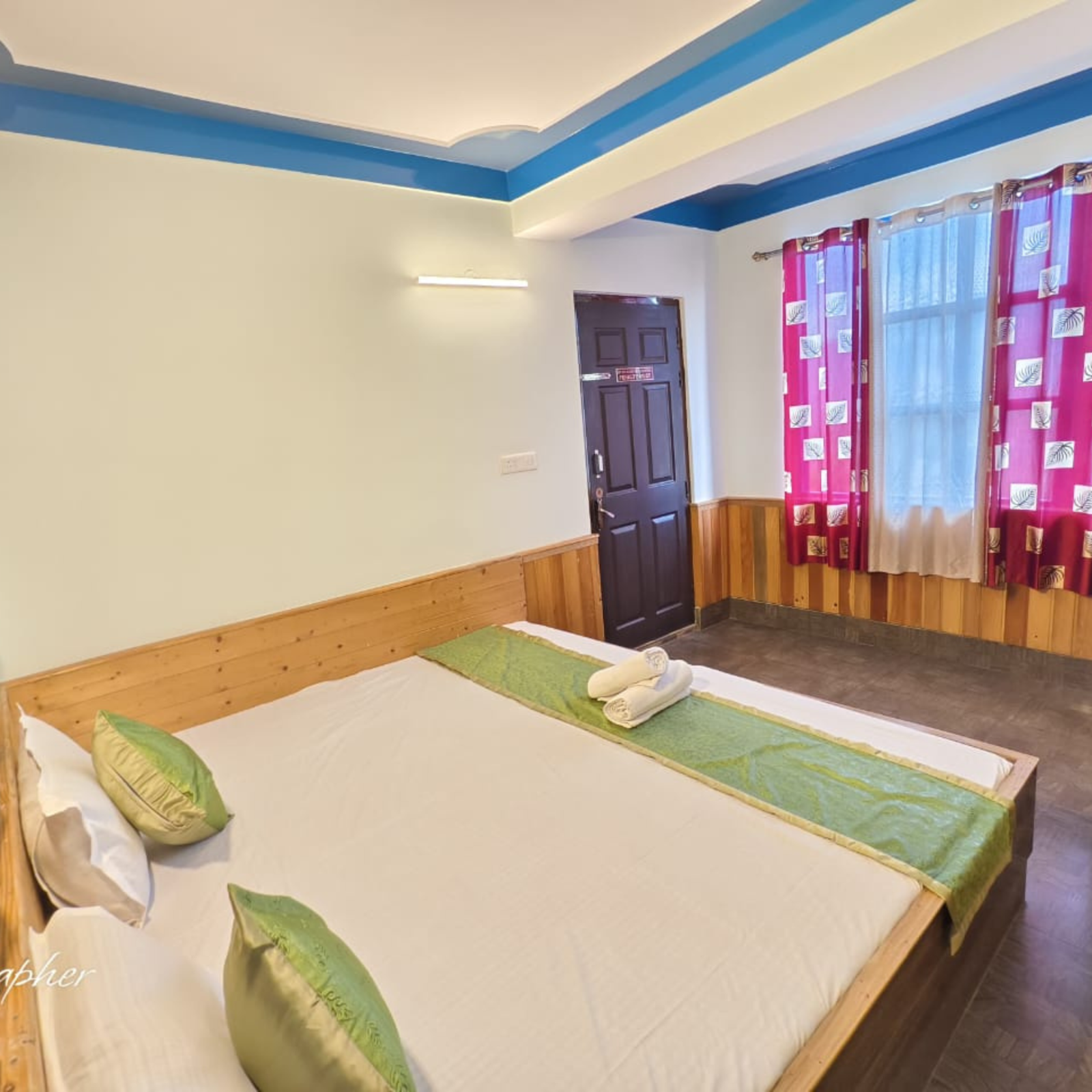 Hotel room with two single beds side by side, white linens, green decorative runners and pillows, wood paneling, and blue ceiling stripes near a window with pink patterned curtains.